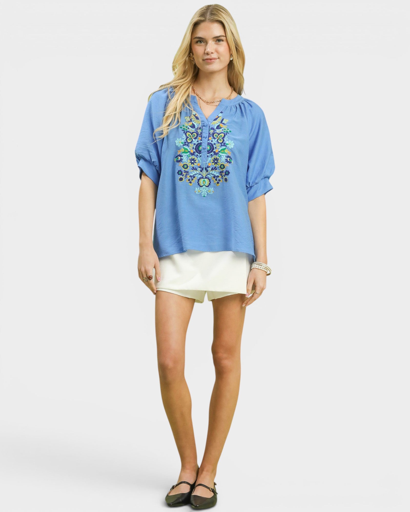 Woman wearing a blue embroidered blouse and white shorts on a plain background