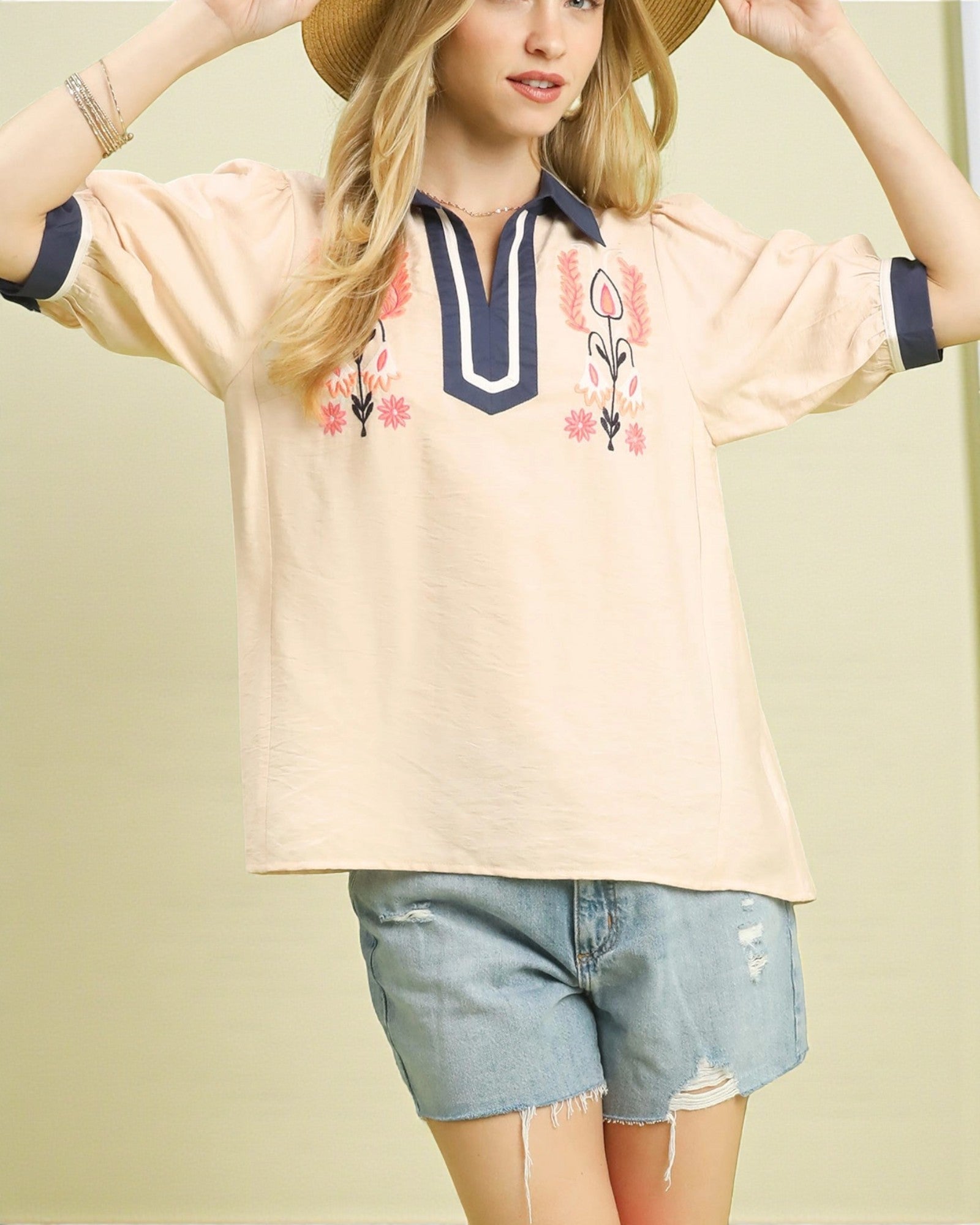 Woman wearing a beige blouse with floral embroidery and denim shorts on a plain background