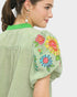 Woman wearing a light green blouse with colorful floral embroidery on a plain background