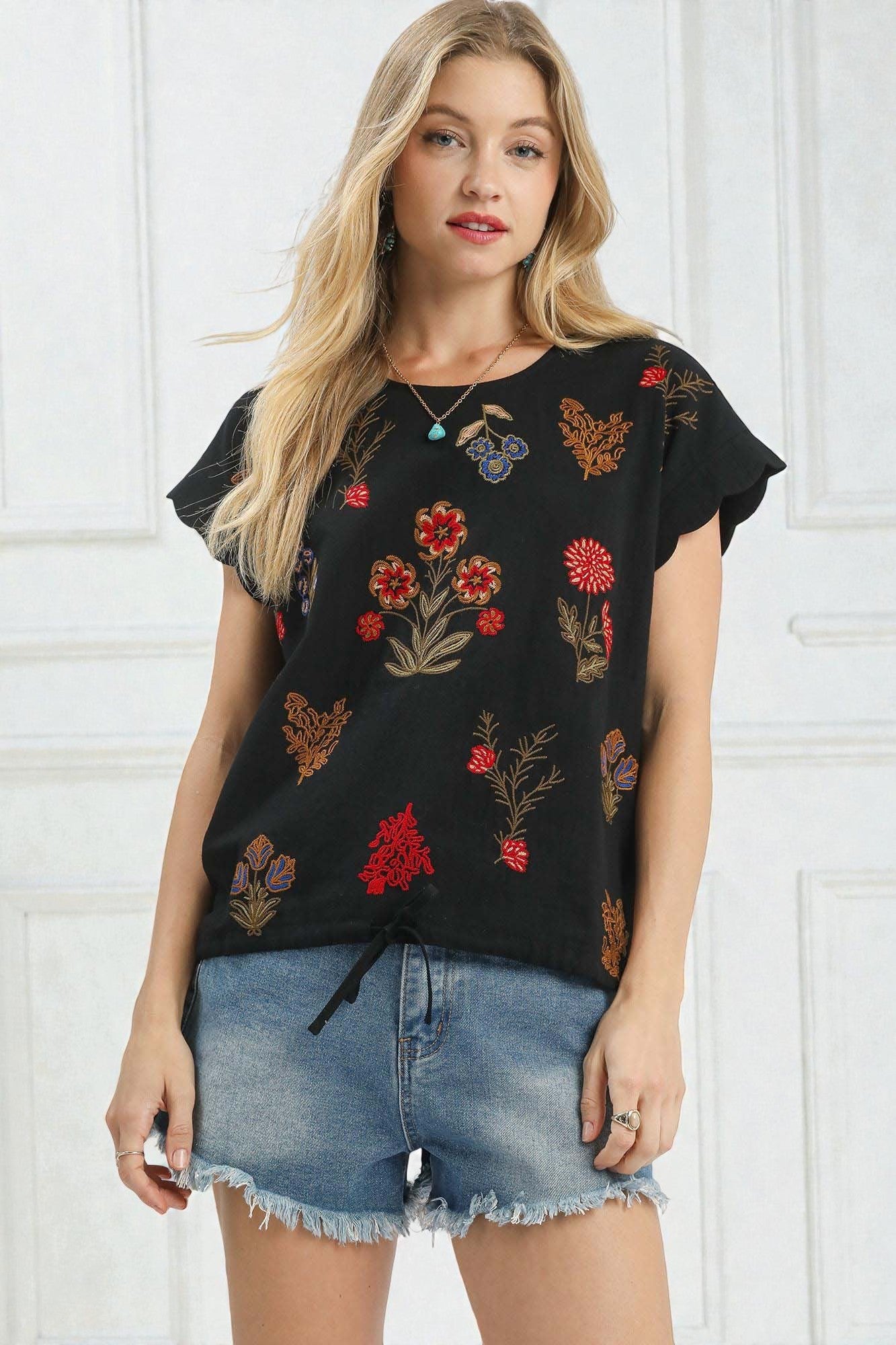 Woman wearing a black floral embroidered top and denim shorts against a white background