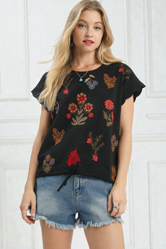 Woman wearing a black floral embroidered top and denim shorts against a white background