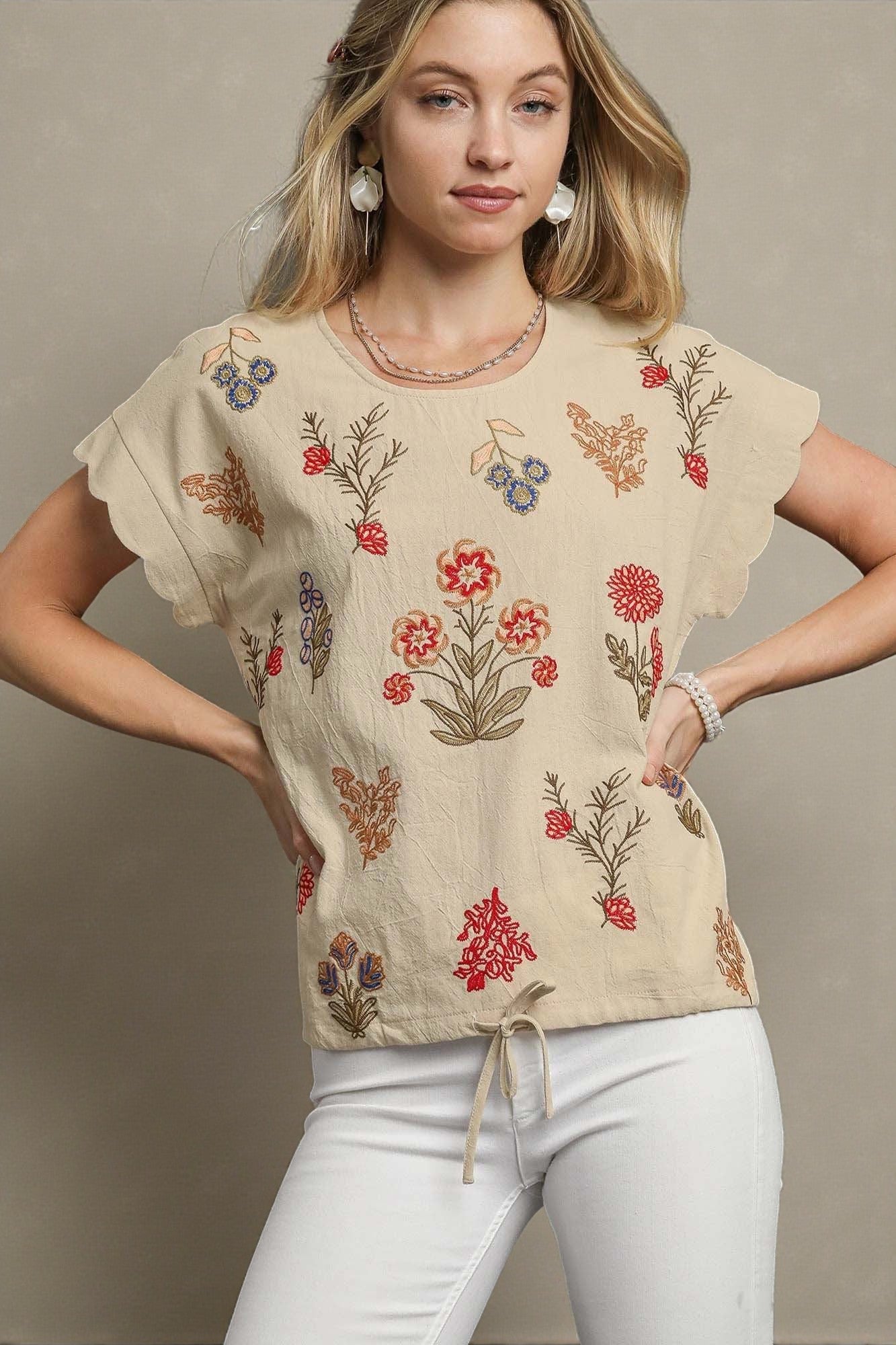 Woman wearing a beige top with floral embroidery against a neutral background
