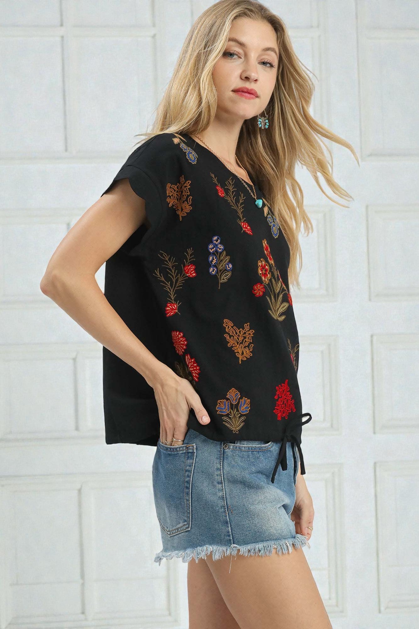 Woman wearing a black top with colorful floral embroidery and denim shorts against a white wall.