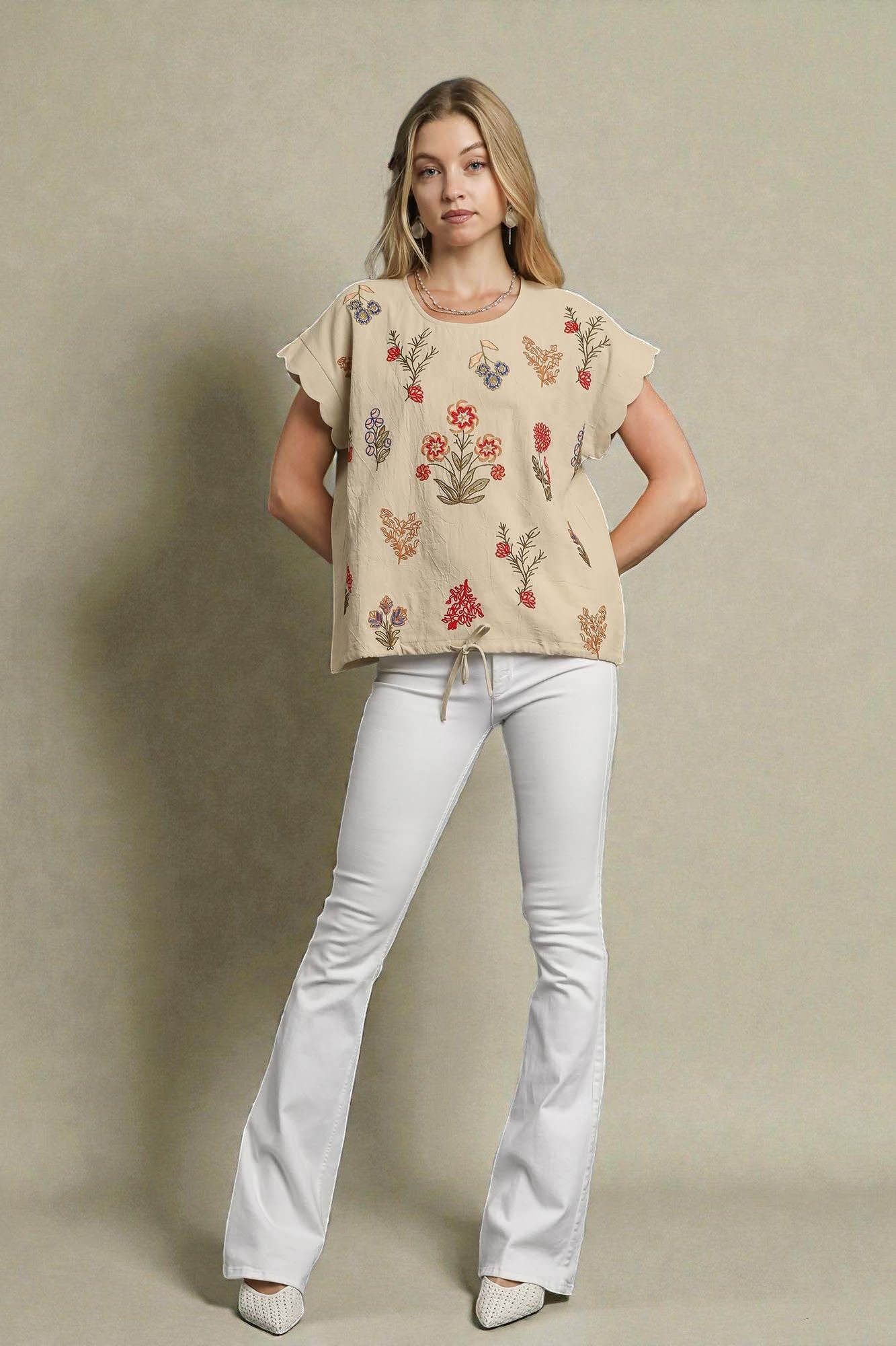 Woman wearing a beige floral embroidered top and white pants against a neutral background