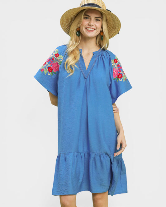 Woman wearing a blue dress with floral embroidery and a straw hat on a white background