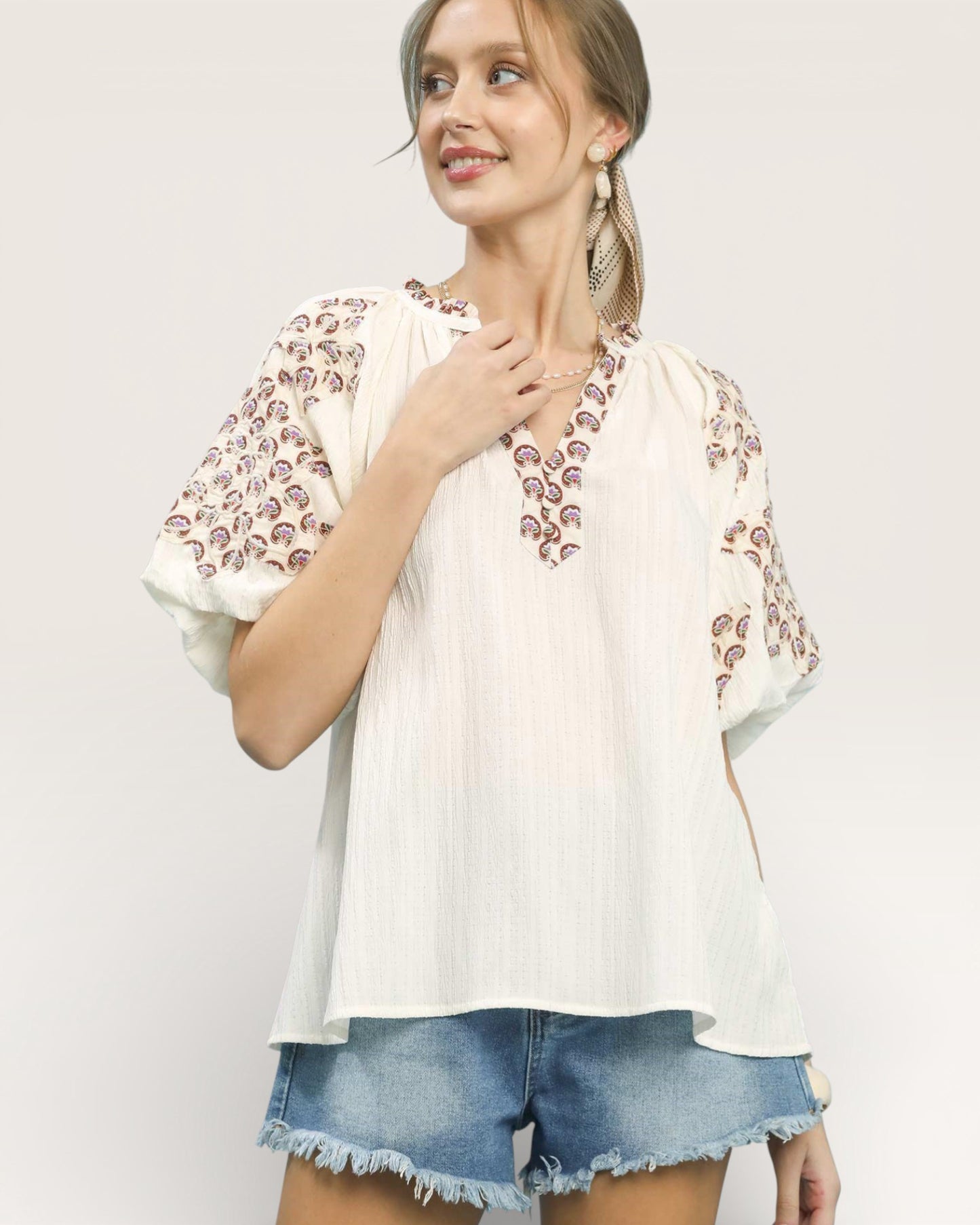 Woman wearing a white blouse with floral embroidery and denim shorts on a light gray background