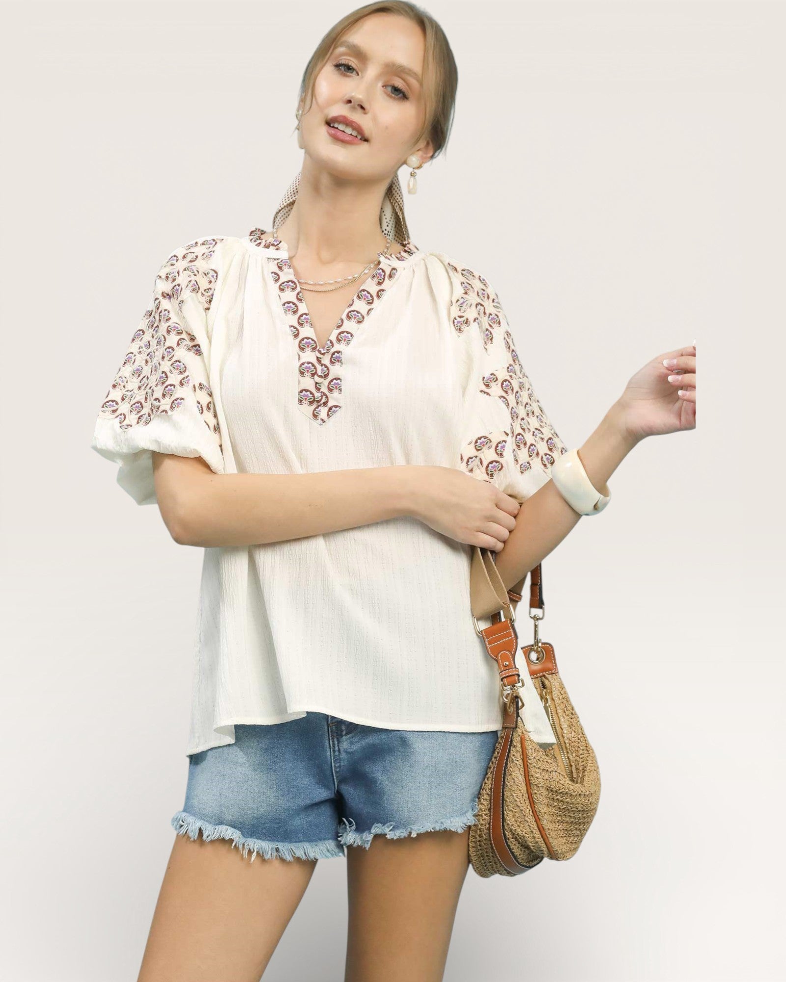 Woman wearing a white blouse with floral embroidery and denim shorts, holding a woven bag on a plain background.