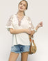 Woman wearing a white blouse with floral embroidery and denim shorts, holding a woven bag on a plain background.