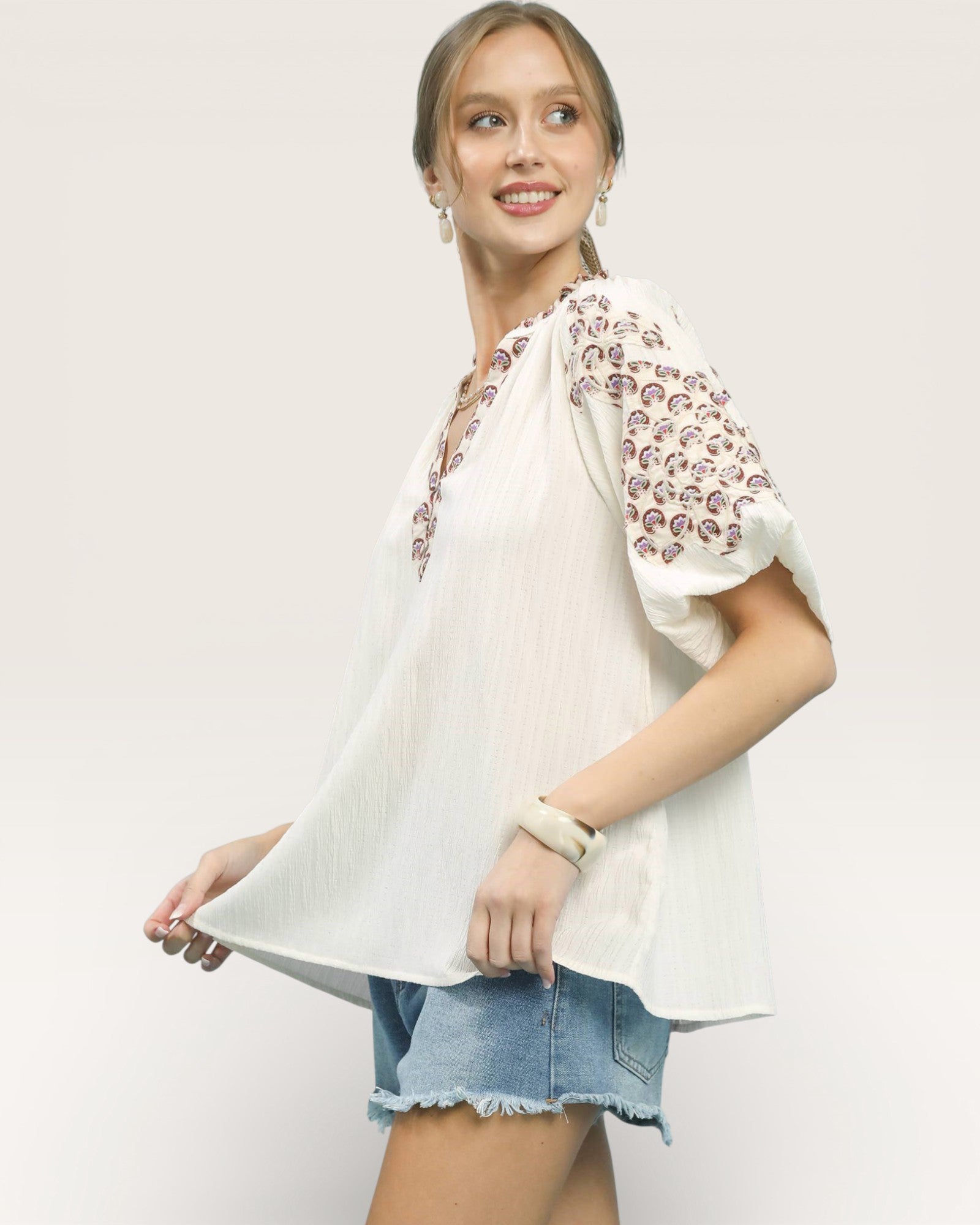 Woman wearing a white blouse with floral sleeves and denim shorts on a plain background