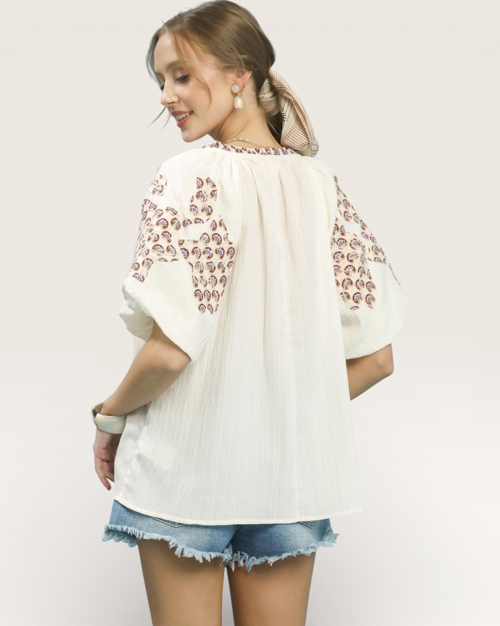 Woman wearing a white blouse with floral embroidery and denim shorts on a plain background