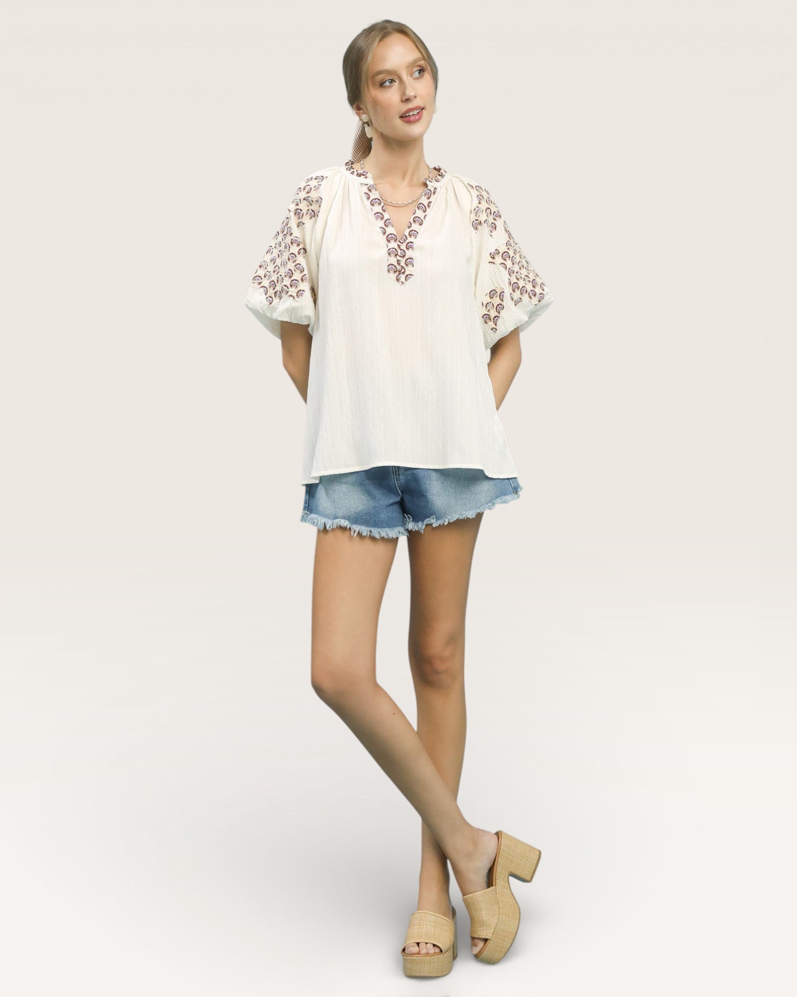 Woman wearing a white blouse with floral embroidery and denim shorts on a plain background