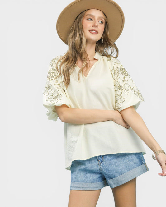 Woman wearing a white blouse with floral embroidery and denim shorts on a plain background