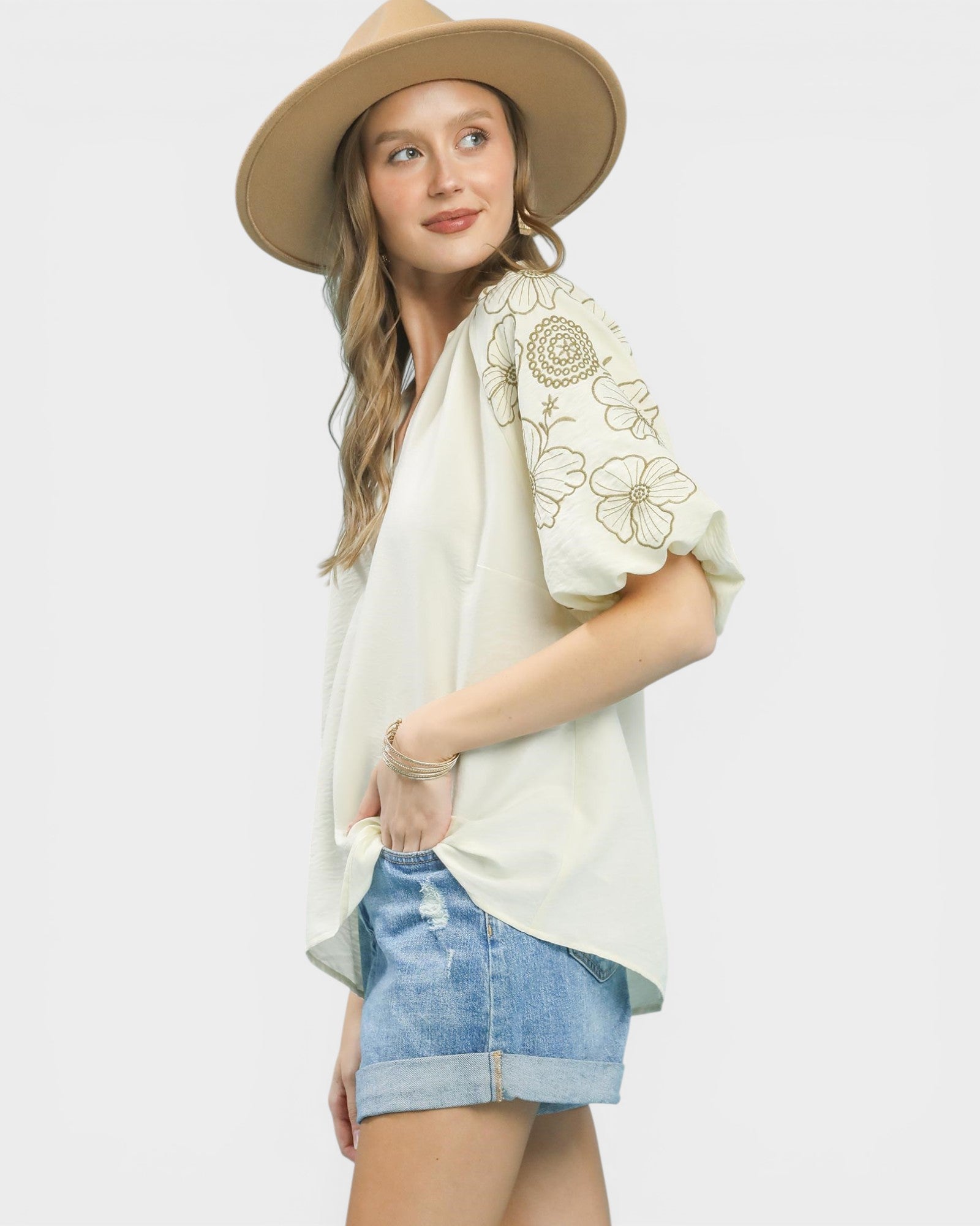 Woman wearing a beige hat, cream blouse with floral embroidery, and denim shorts on a white background