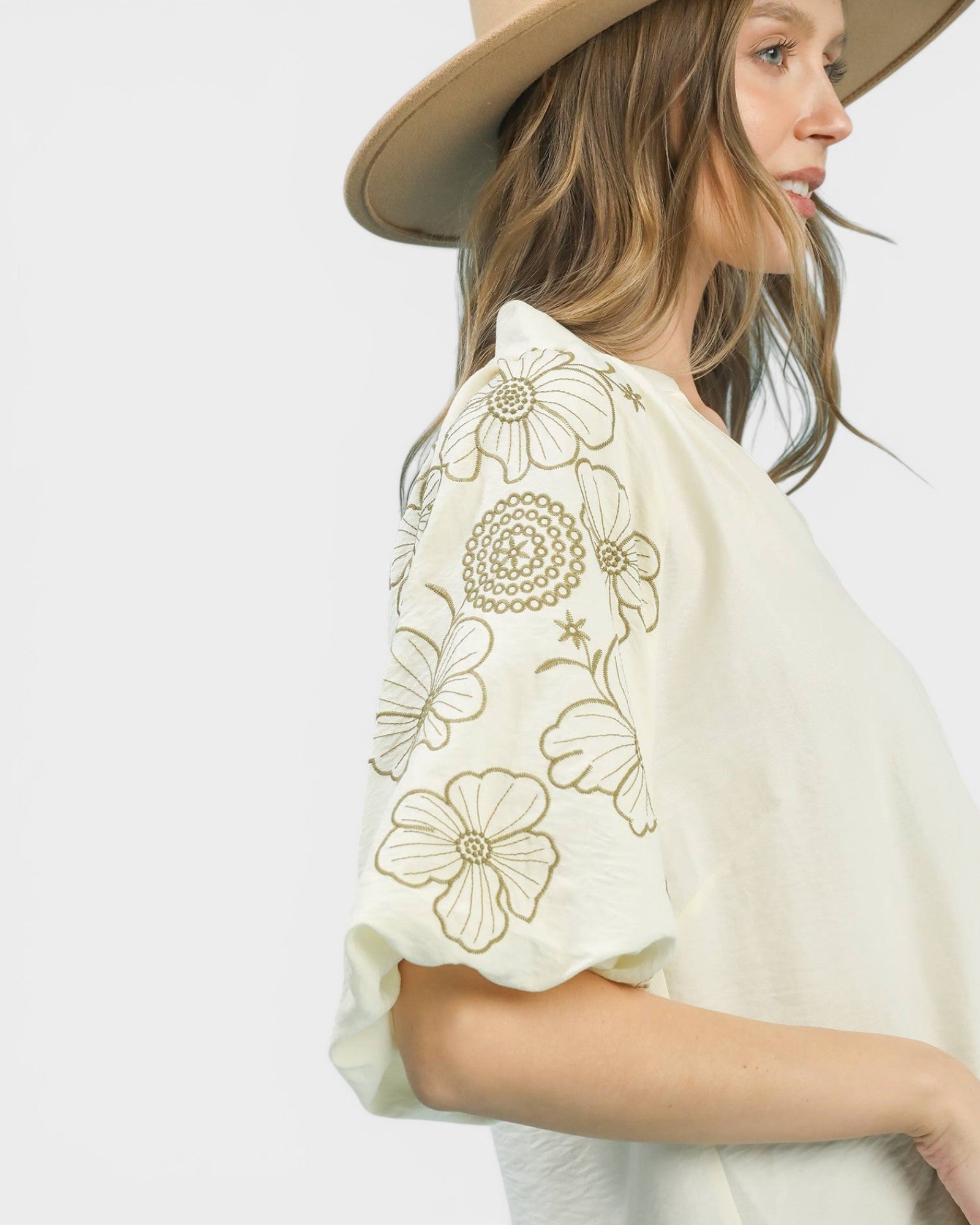 Woman wearing a white blouse with floral embroidery and a beige hat on a light gray background
