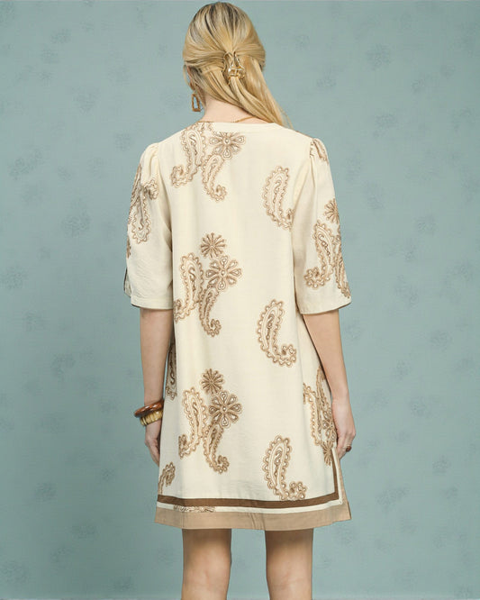 Woman wearing a cream dress with gold embroidery against a light blue background