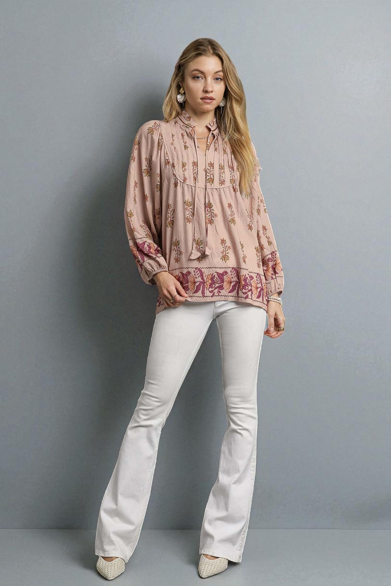 Woman wearing a patterned blouse and white pants against a gray background