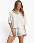 A woman modeling a matching two-piece lounge set in a textured off-white fabric, featuring a short-sleeve collared top and coordinating shorts, set against a plain white studio background.