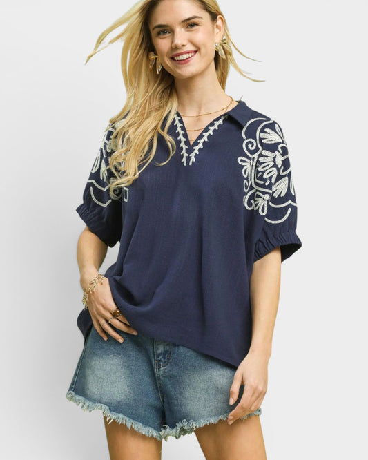 Woman wearing a navy blue blouse with white embroidery and denim shorts on a light gray background