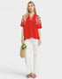 Woman wearing a red blouse with white embroidery and white pants, holding a woven bag with plants.