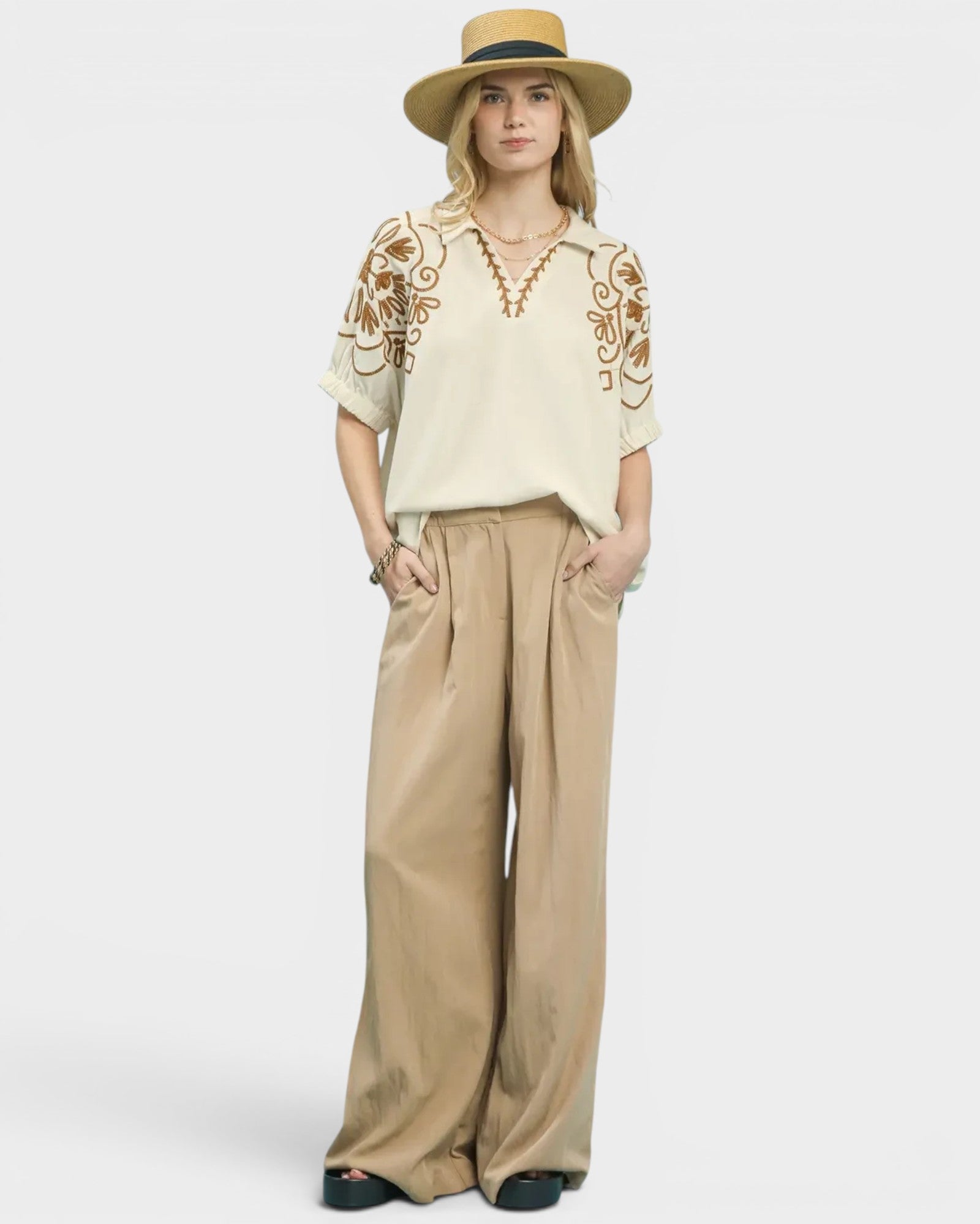 Woman wearing a cream embroidered top and beige wide-leg pants with a hat on a white background