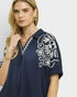Woman wearing a navy blue top with white floral embroidery on a plain background