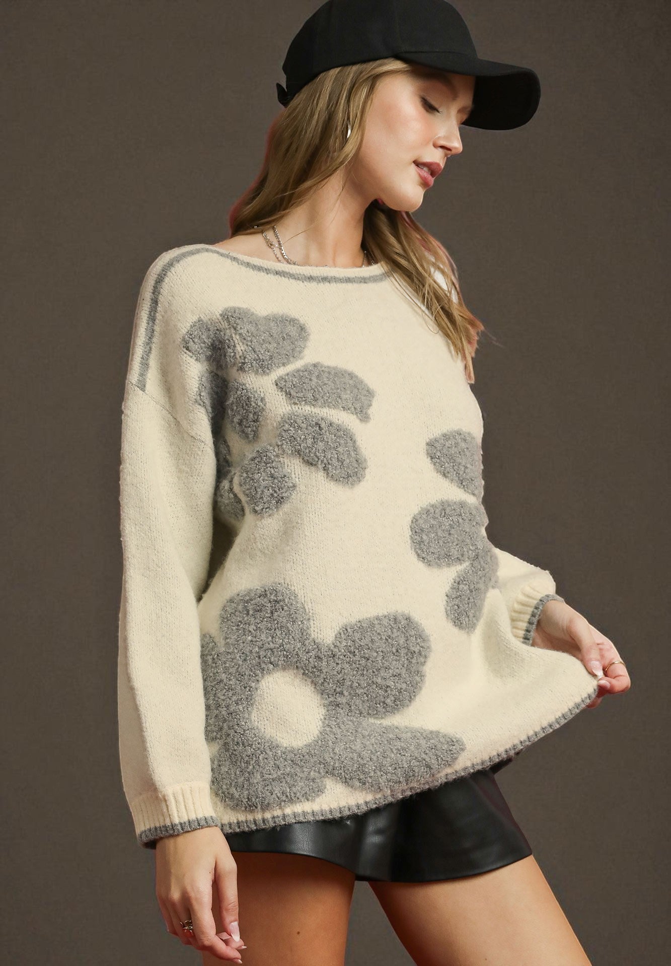 Woman wearing a cream sweater with gray floral patterns and black cap on a brown background