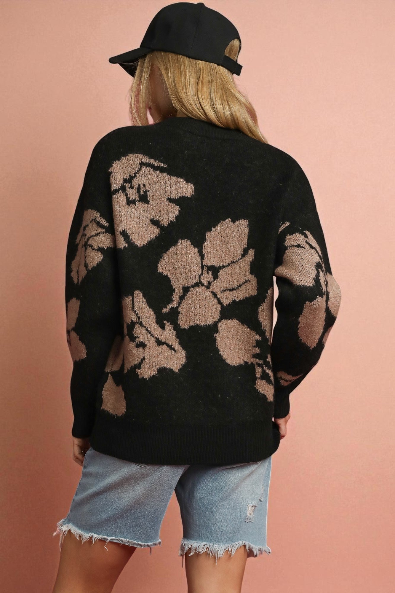 Person wearing a black floral-patterned sweater and denim shorts against a pink background