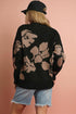 Person wearing a black floral-patterned sweater and denim shorts against a pink background
