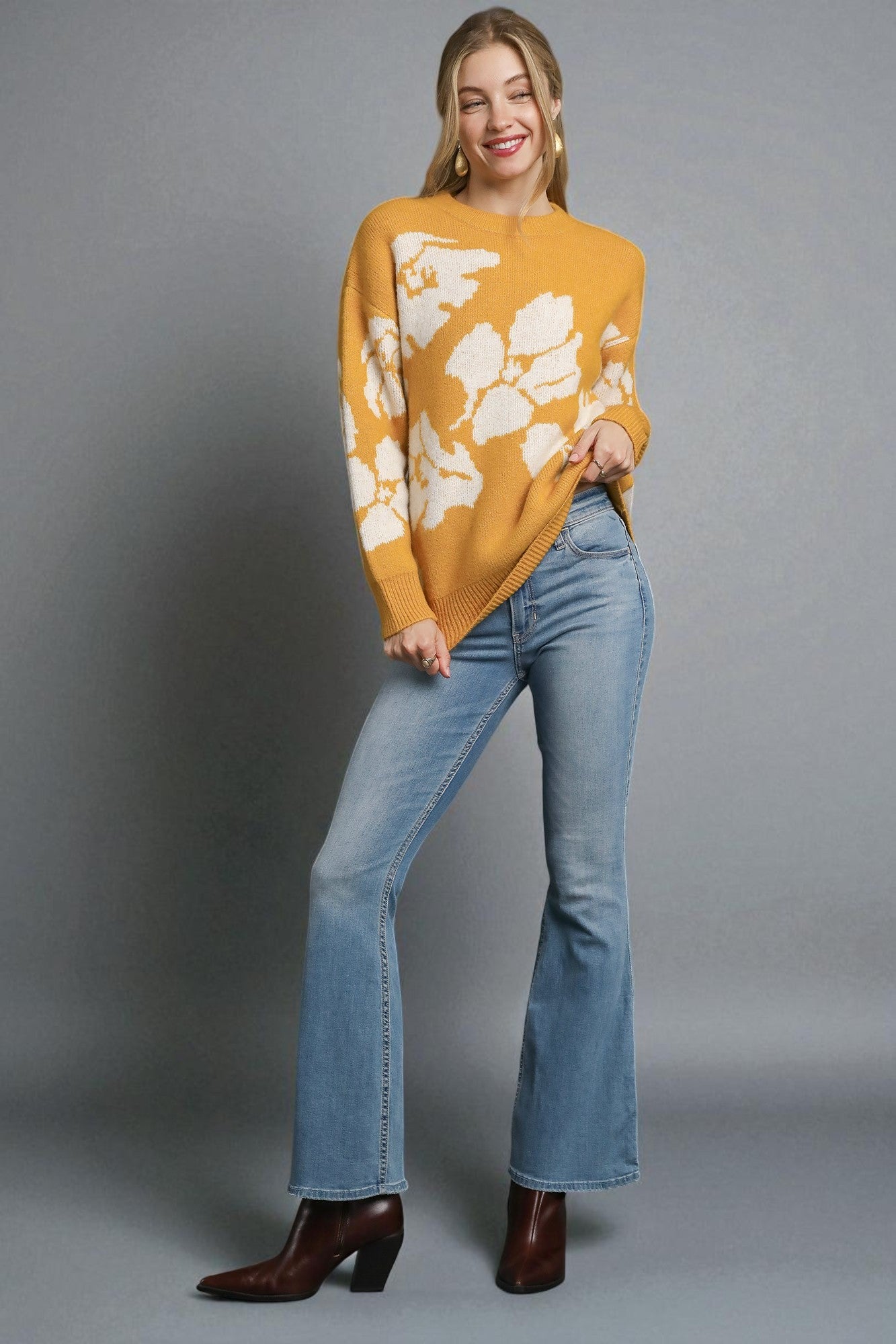 Woman wearing a mustard yellow sweater with white floral patterns and blue jeans on a gray background