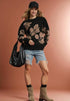 Woman wearing a black floral sweater, denim shorts, and brown boots against a pink background