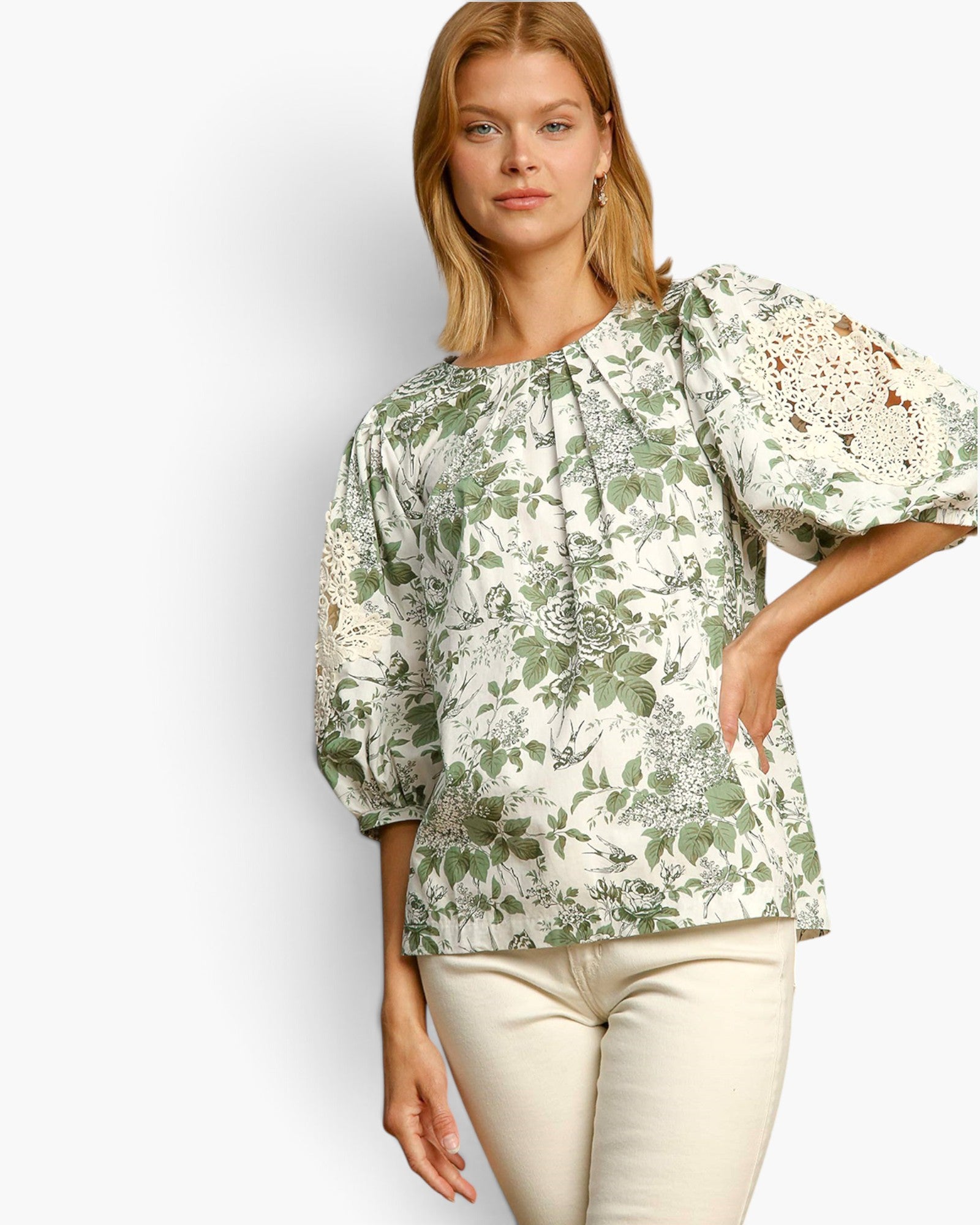 A woman modeling a cream-colored blouse with a green botanical and floral print, featuring puff sleeves with lace applique on the upper arms, paired with cream pants against a light gray studio background.