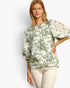 A woman modeling a cream-colored blouse with a green botanical and floral print, featuring puff sleeves with lace applique on the upper arms, paired with cream pants against a light gray studio background.