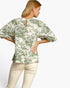 A woman shown from the back wearing a cream-colored blouse with a green floral pattern and puff sleeves, paired with light beige pants, against a plain white background.