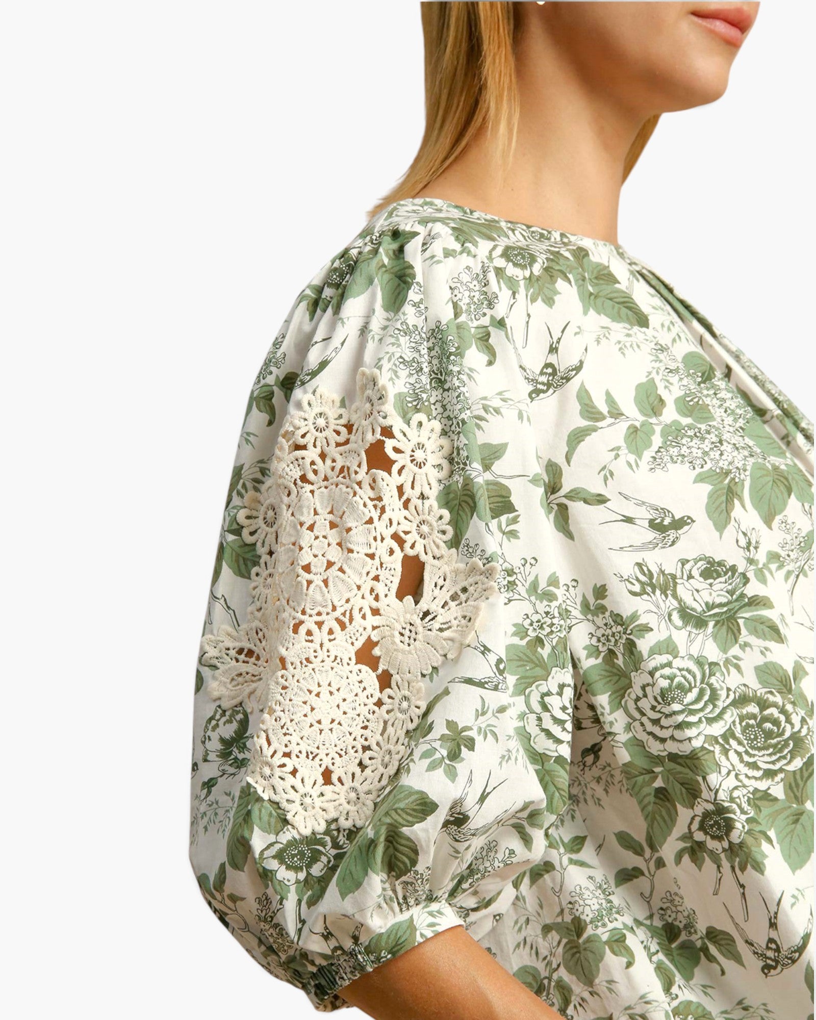 A close-up side view of a woman wearing a cream blouse with a green floral and bird print, featuring eyelet lace puff sleeves.