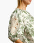 A close-up side view of a woman wearing a cream blouse with a green floral and bird print, featuring eyelet lace puff sleeves.