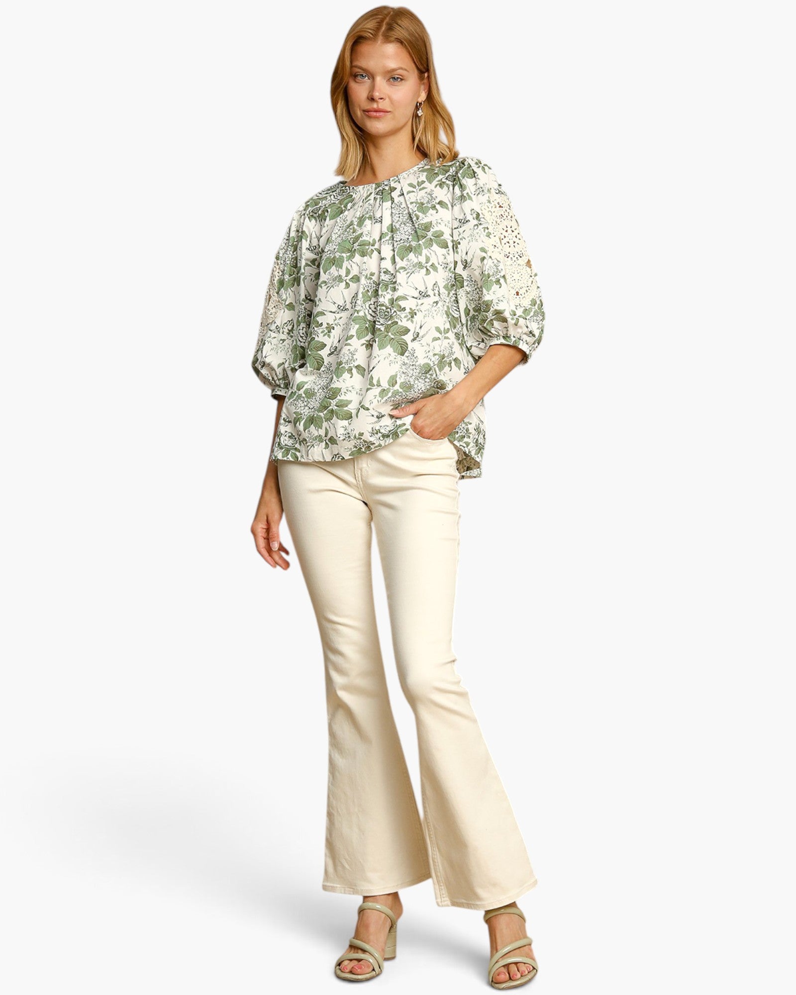A woman modeling a loose, short-sleeved blouse with a green and white floral print, paired with cream-colored flared pants and metallic sandals, set against a plain light gray studio background.