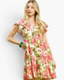 A woman modeling a short-sleeve tiered dress with a vibrant pink, red, and green floral print on a cream background, set against a plain light gray studio backdrop.