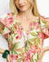 A close-up of a woman wearing a cream-colored dress with a vibrant pink, red, and green floral print, featuring ruffled sleeves and a V-neckline.