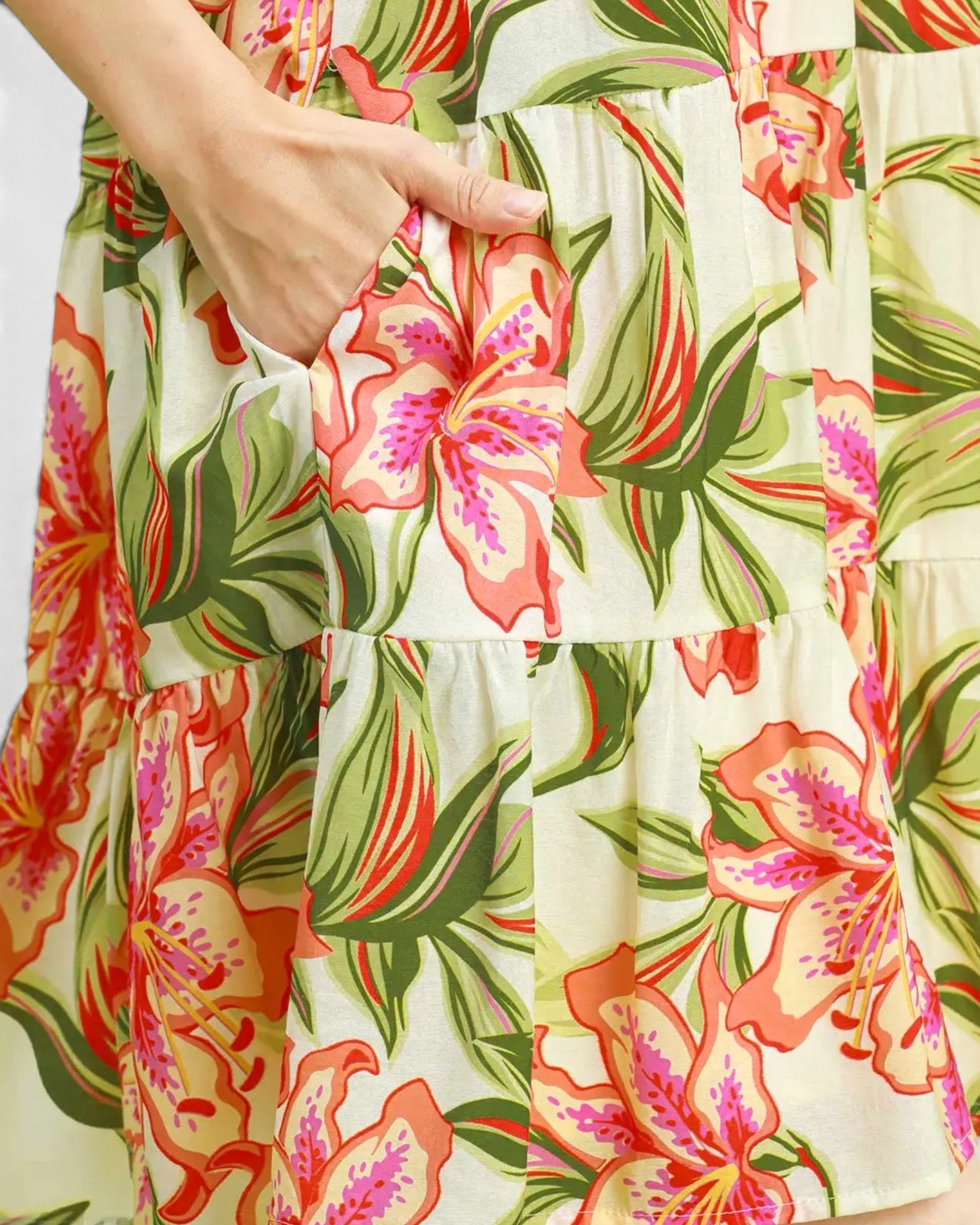 Close-up of a tiered dress with a vibrant tropical floral print, featuring large pink and orange flowers with green leaves on an off-white background. A hand is shown reaching into a side pocket.