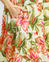 Close-up of a tiered dress with a vibrant tropical floral print, featuring large pink and orange flowers with green leaves on an off-white background. A hand is shown reaching into a side pocket.