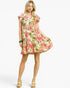 A woman modeling a short tiered dress with a pink and green tropical floral print on a cream background, paired with gold sandals and a green bracelet, against a light gray studio background.