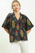 Woman wearing a black floral blouse with white pants on a plain background