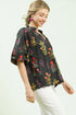 Woman wearing a black floral blouse with white pants on a light background