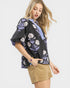 Woman wearing a black floral blouse with purple accents and beige shorts on a white background