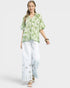 Woman wearing a green patterned top and white pants on a light gray background