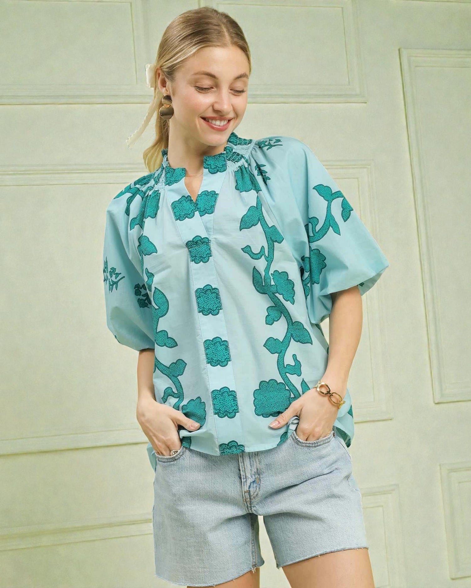 Woman wearing a light blue blouse with green floral patterns against a light green wall.