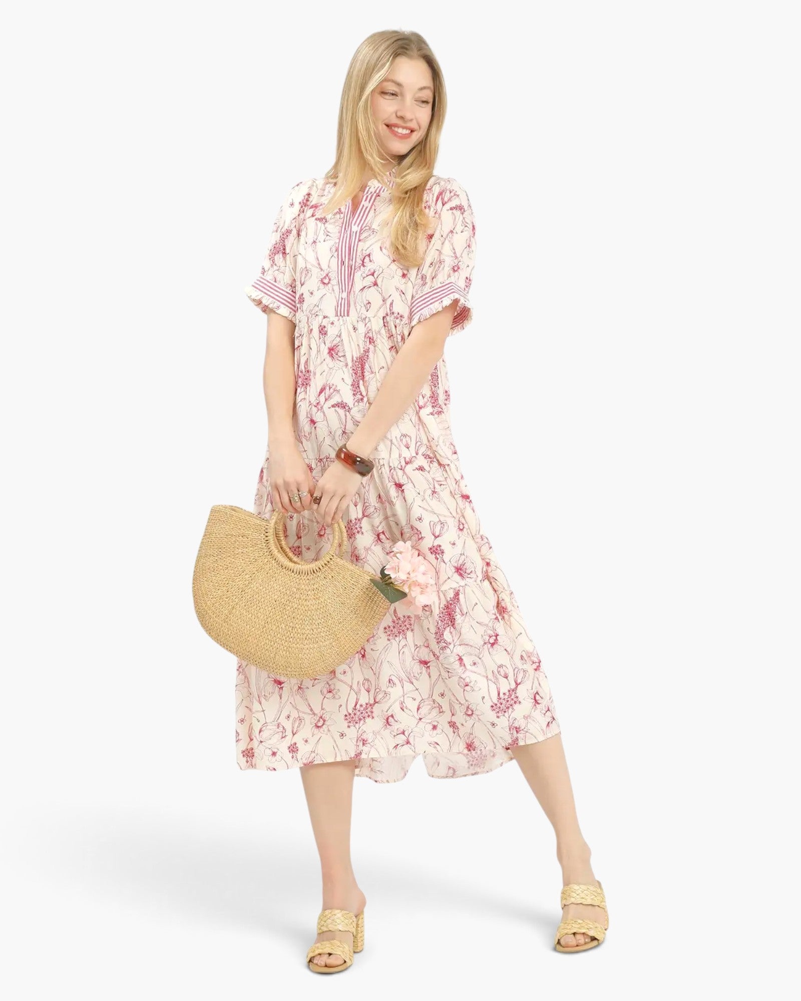 A woman modeling a short-sleeve midi dress with a pink and red abstract floral print on a cream background, holding a woven straw handbag and wearing gold slide sandals against a light gray studio background.