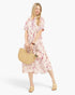 A woman modeling a short-sleeve midi dress with a pink and red abstract floral print on a cream background, holding a woven straw handbag and wearing gold slide sandals against a light gray studio background.