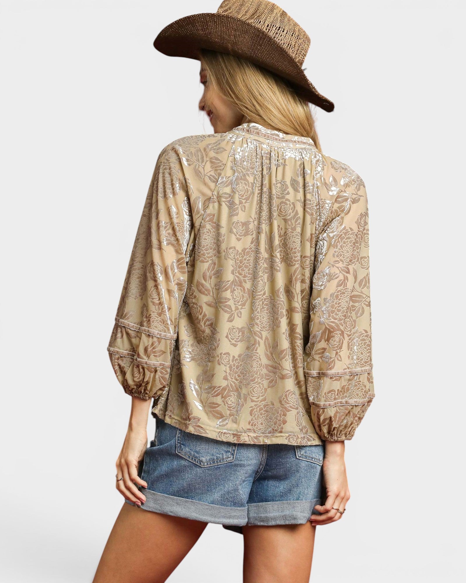 Woman wearing a beige patterned blouse and denim shorts with a brown hat on a white background