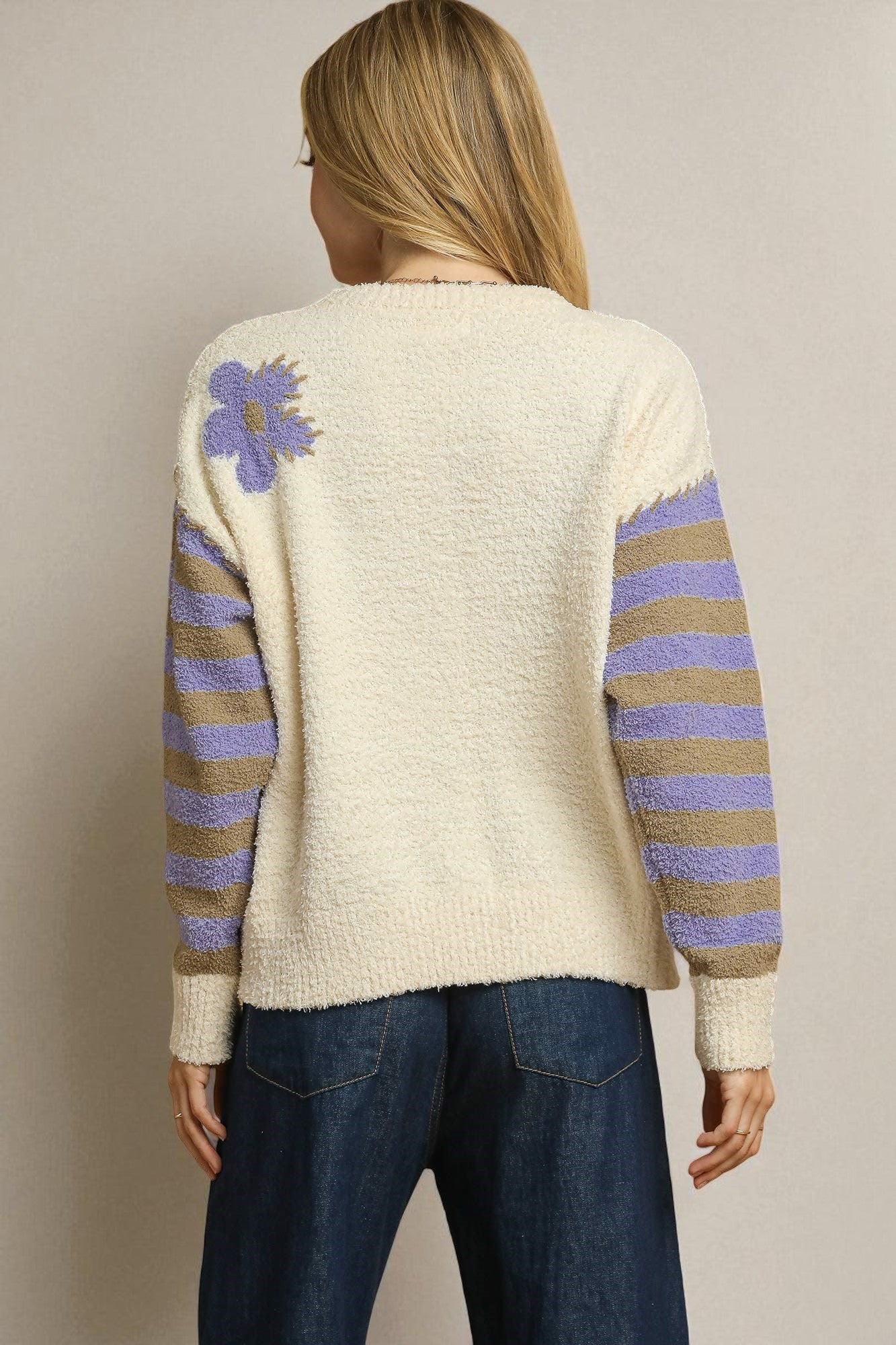 Person wearing a cream sweater with purple and brown stripes and a floral design on the back, against a beige background.